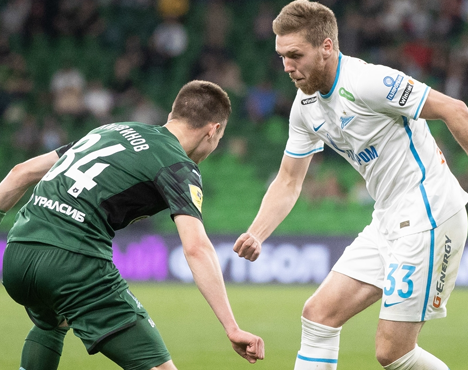 На изображении два футболиста на поле: один в белой форме, другой в зелёной. Они активно взаимодействуют, возможно, борются за мяч. На фоне видны размытия зрителей трибун